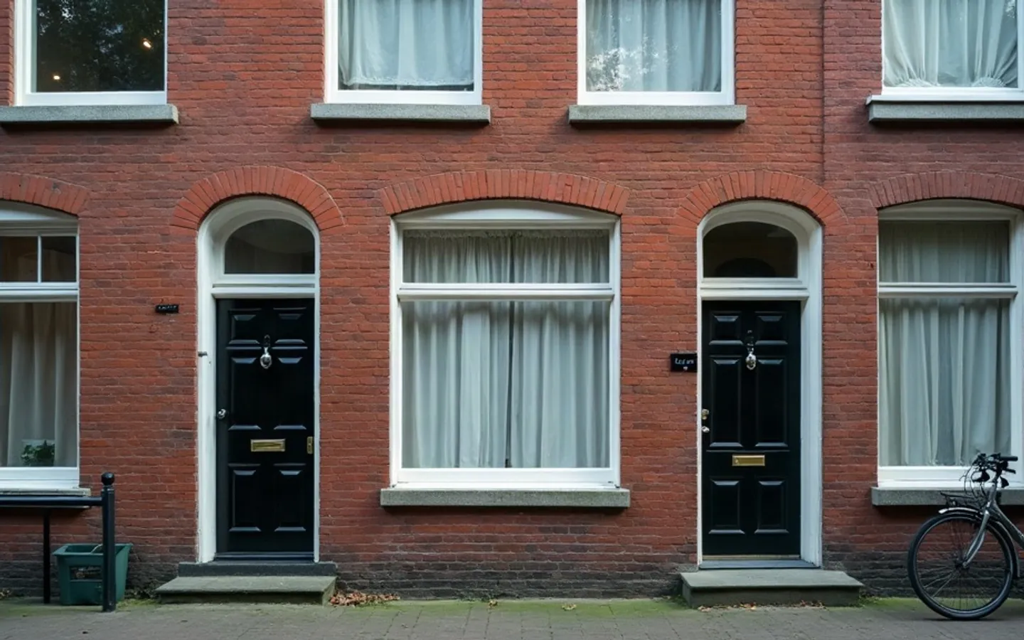 Bakstenen gevel van twee Nederlandse benedenwoningen met zwarte voordeuren, gesloten vitrages en een fiets tegen de gevel.